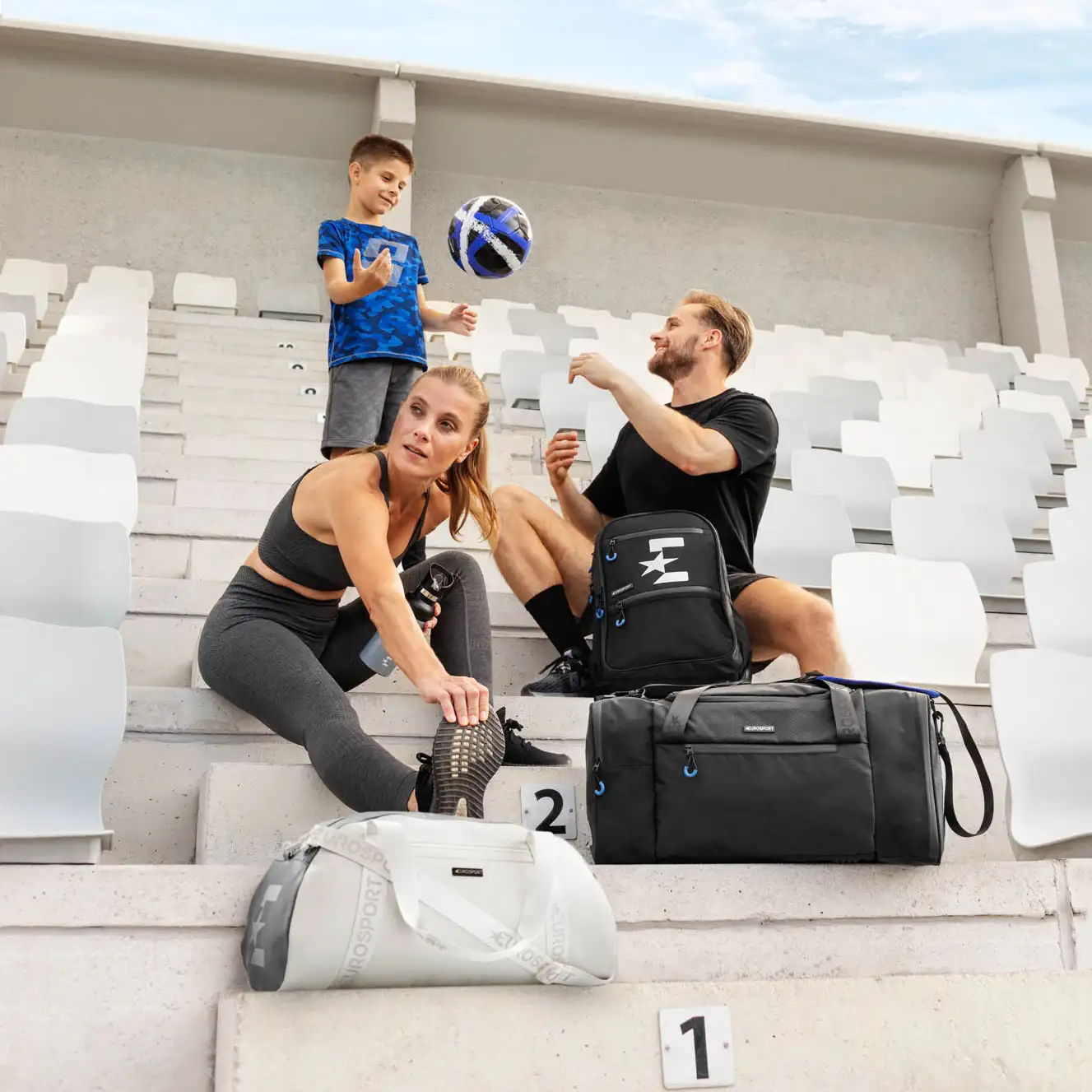 Športna družina na tribuni s torbami Eurosport – priprava na trening in raztezanje v sproščenem ambientu.